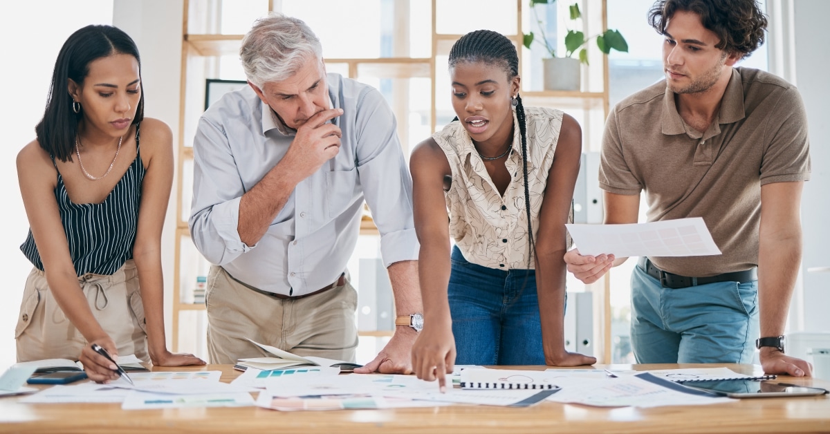 Four team members standing and working together at a table. One woman is pointing at a document on the table and everyone is looking at it. The image is used as the featured image for the "How Can My Staffing Firm Sell More in Today's Economy?" blog post.